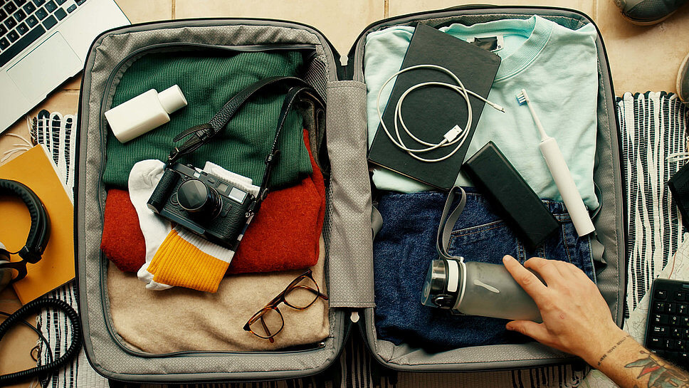Mann packt offenen Koffer mit Kleidung, elektronischen Geräten und Kosmetik
