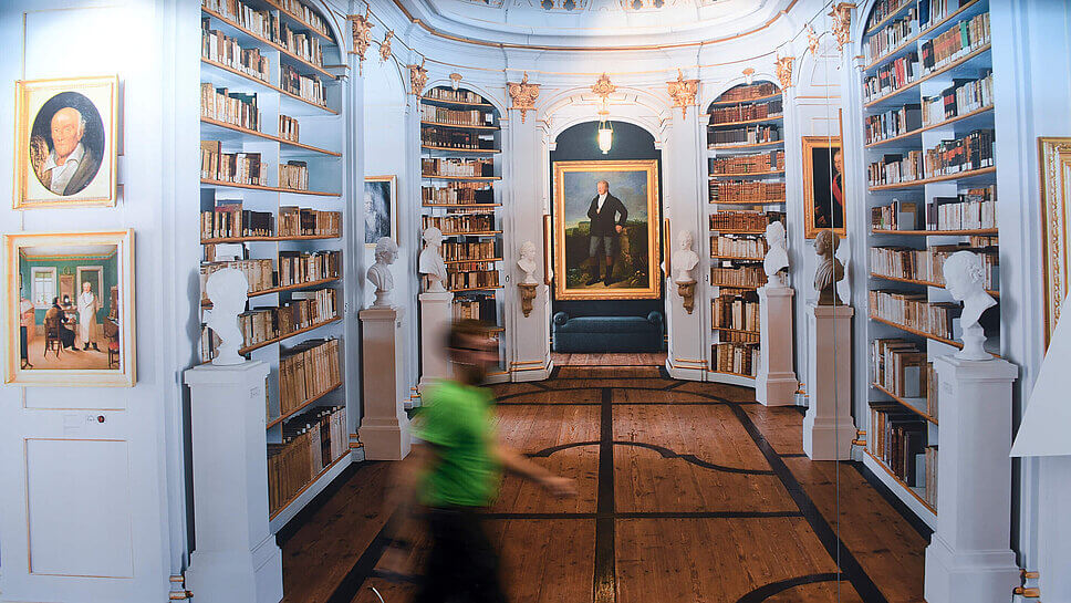 Eine Person, undeutlich zu erkennen, durchschreitet einen Bibliotheksraum mit dicht gefüllten Buchregalen 