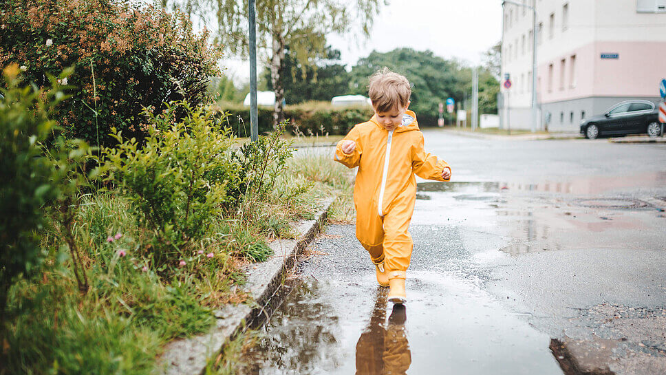 Ein Junge im gelben Regenanzug läuft durch eine Pfütze, Büsche am Straßenrand