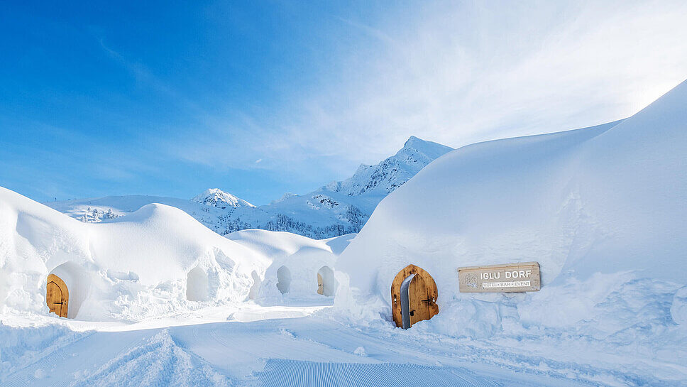 Aus Schnee gebaute Iglus mit Holztüren, verschneite Wege, Bergpanorama im Hintergrund