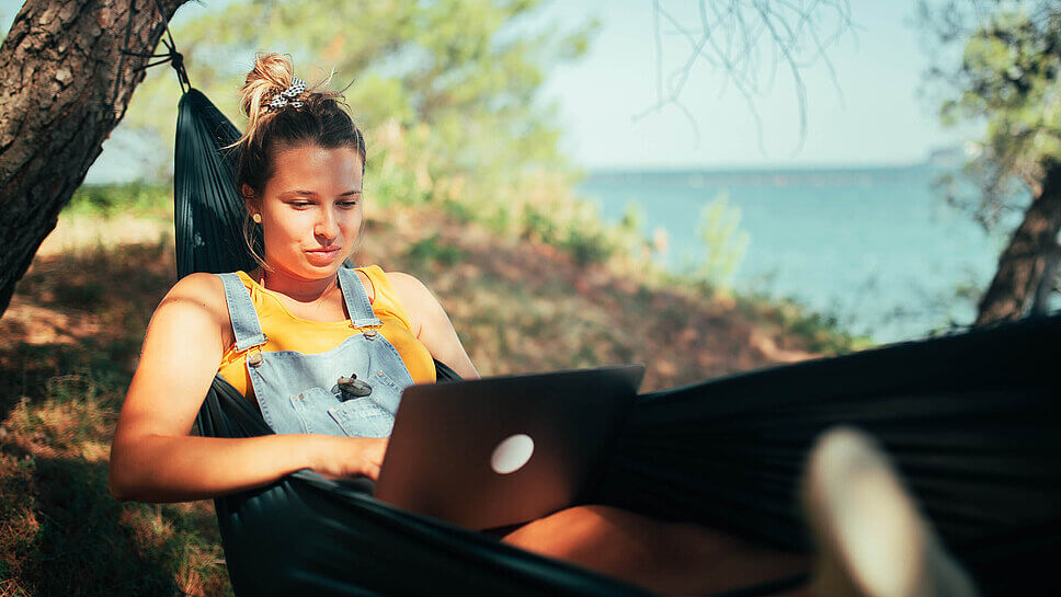 Eine Frau liegt an einem sonnigen Ort mit Laptop in der Hängematte und arbeitet 