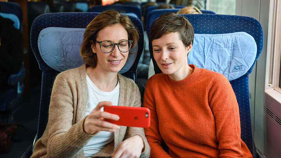 Zwei Frauen blicken in der Bahn auf ein Handy, sitzend