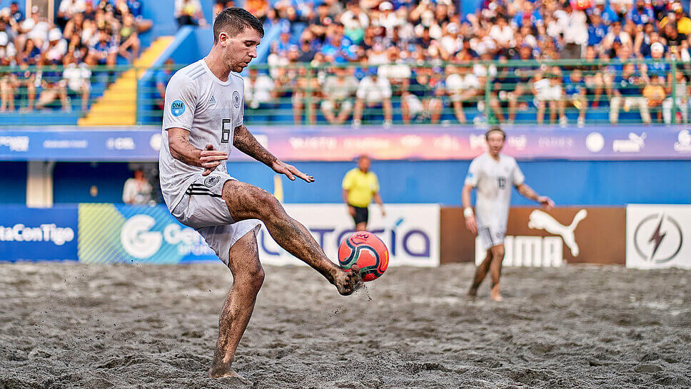 Mann lupft einen Fußball mit dem Fuß, er spielt auf Sand, im Hintergrund eine Tribüne